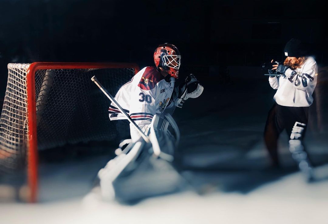 Hockey goalie in full gear stands ready on the ice, another person films him on ice rink with dark background, spotlighting the subjects.