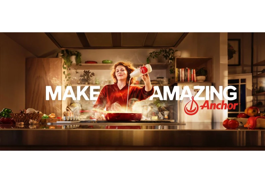Woman in a kitchen pouring milk into a red pan, with a logo for Anchor, indoor kitchen setting, with smoke, cooking ingredients on the counter.