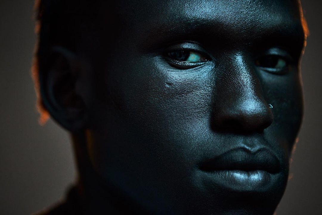 Striking close-up studio portrait of a dark-skinned adult male with visible facial textures, a nose ring, and dramatic rim lighting.