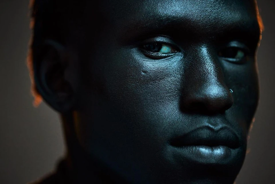 Striking close-up studio portrait of a dark-skinned adult male with visible facial textures, a nose ring, and dramatic rim lighting.