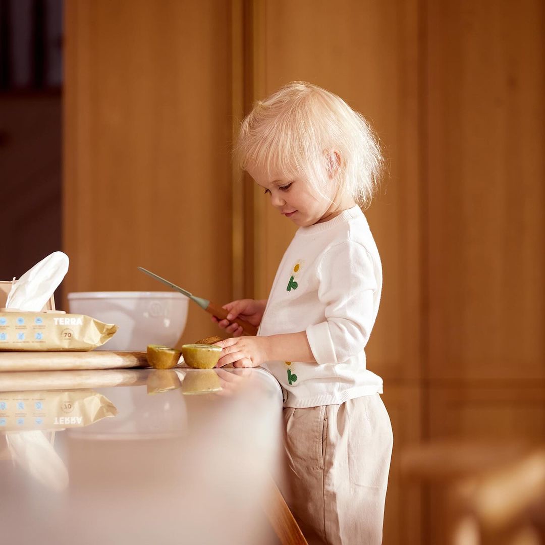 A young child with blonde hair is slicing a halved kiwi fruit on a kitchen countertop with a knife, set against a softly lit interior.