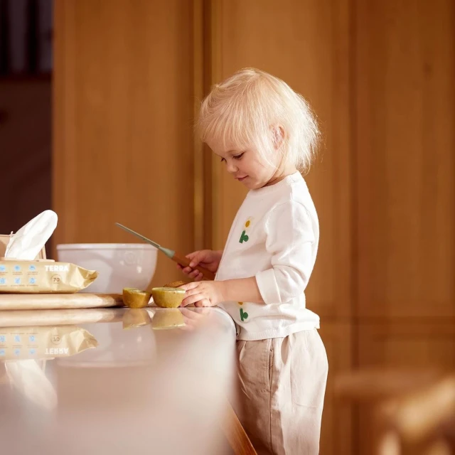 A young child with blonde hair is slicing a halved kiwi fruit on a kitchen countertop with a knife, set against a softly lit interior.