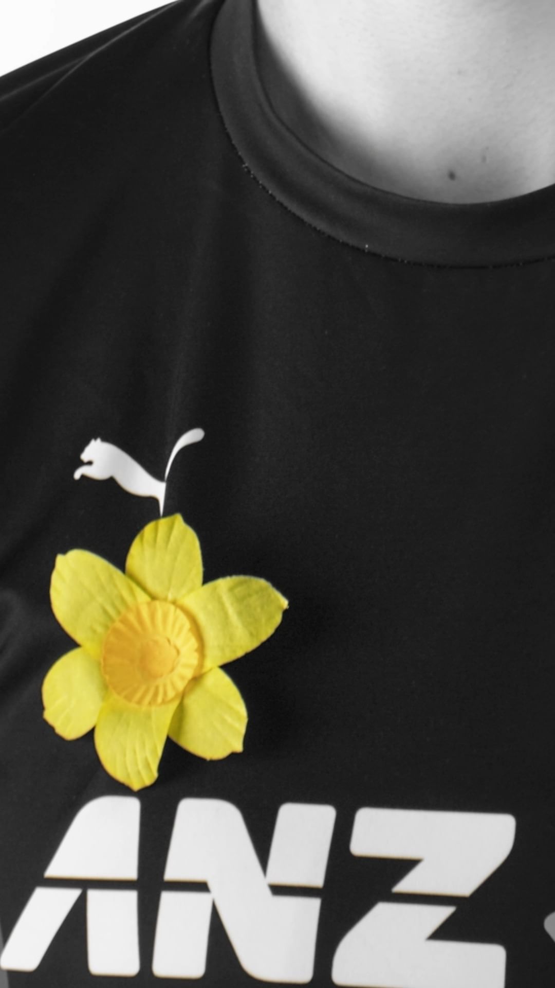 Close-up of a black t-shirt with a yellow daffodil pinned on it, featuring the white Puma logo and the white 'ANZ' text logo.