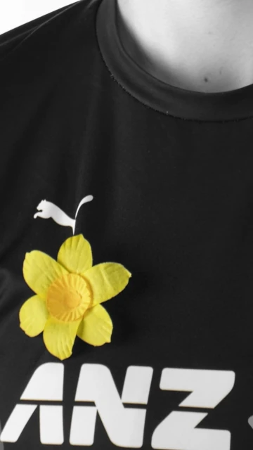 Close-up of a black t-shirt with a yellow daffodil pinned on it, featuring the white Puma logo and the white 'ANZ' text logo.