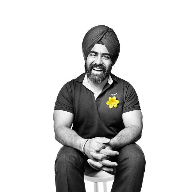 Man in turban smiles seated on stool, in a black and white portrait with a yellow flower on his dark polo shirt against white.