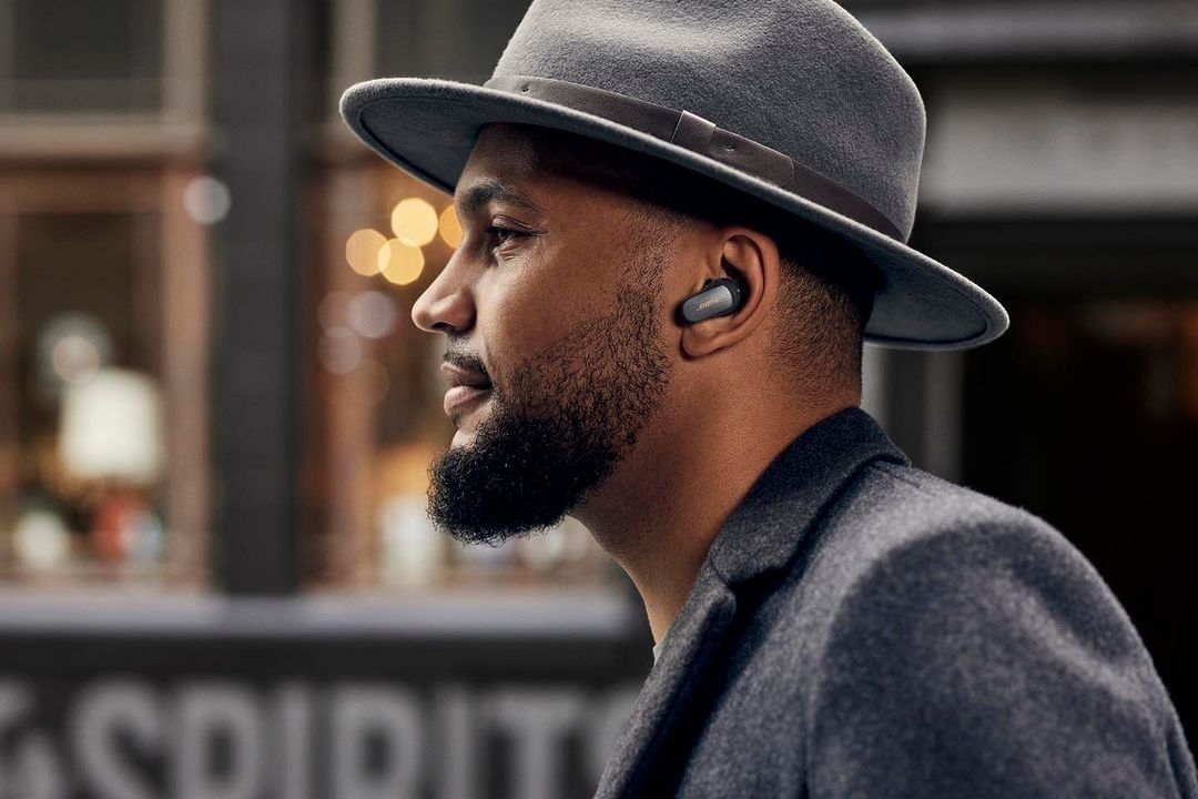 Profile shot of an adult male wearing gray hat and jacket, and earbuds, in an urban environment with blurred building background.