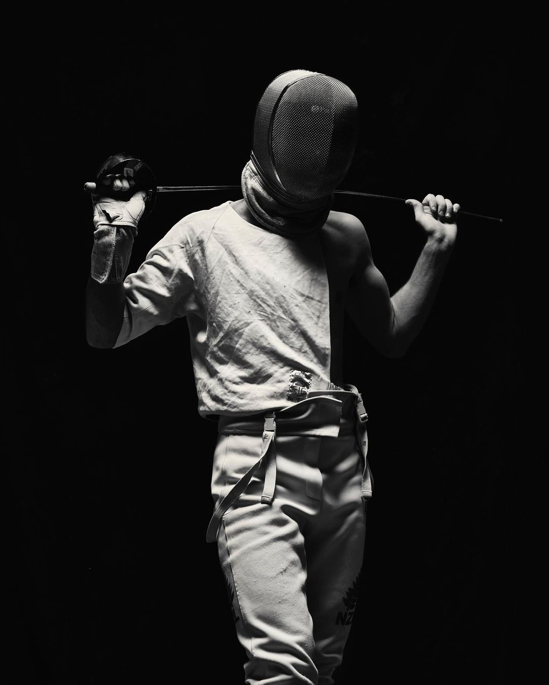 Monochrome portrait of a fencer in uniform with mask, holding a foil, posing against a dark background in a dramatic, high-contrast light.
