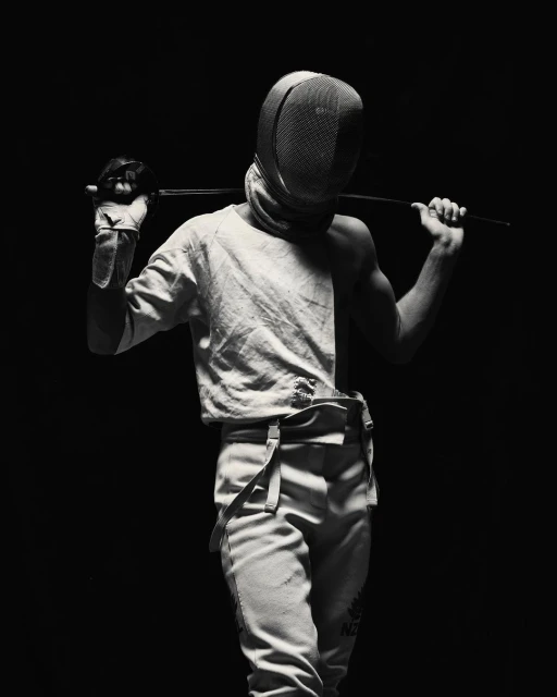 Monochrome portrait of a fencer in uniform with mask, holding a foil, posing against a dark background in a dramatic, high-contrast light.
