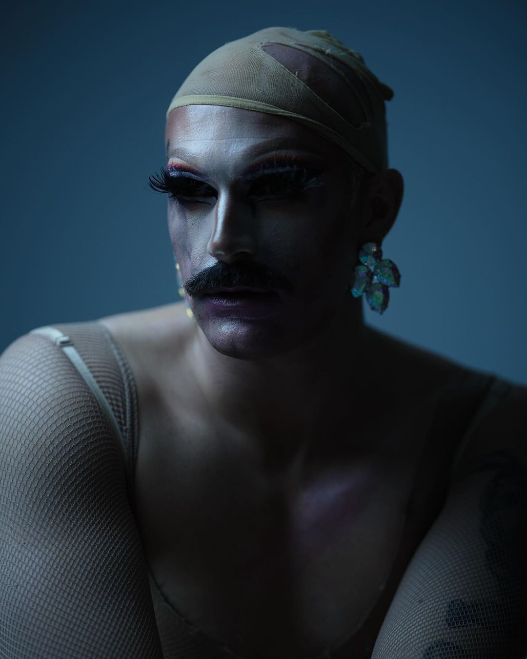 Portrait of a person with a moustache wearing theatrical makeup and an earring, standing indoors with a plain background and dramatic lighting.