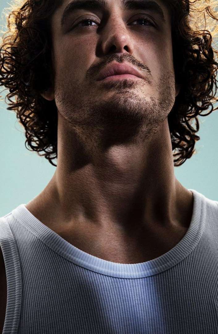 Close-up portrait of a light-skinned adult male with curly hair, wearing a white tank top against a light blue studio backdrop.