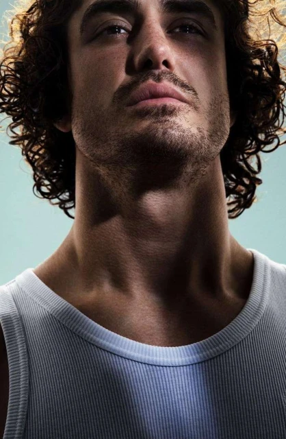 Close-up portrait of a light-skinned adult male with curly hair, wearing a white tank top against a light blue studio backdrop.