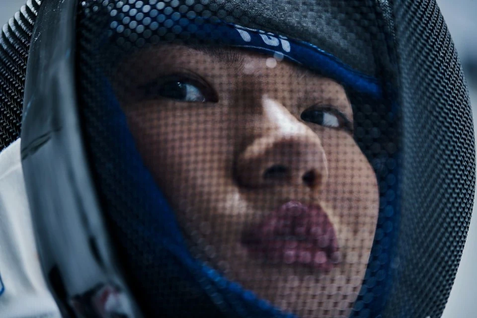 Close up of a female fencer wearing a mesh helmet and bandana, eyes looking up, indoors with soft lighting and shallow depth of field.