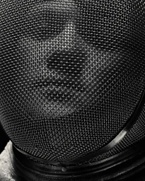 Close-up portrait of a person's face obscured behind a fine wire mesh fencing mask in black and white.