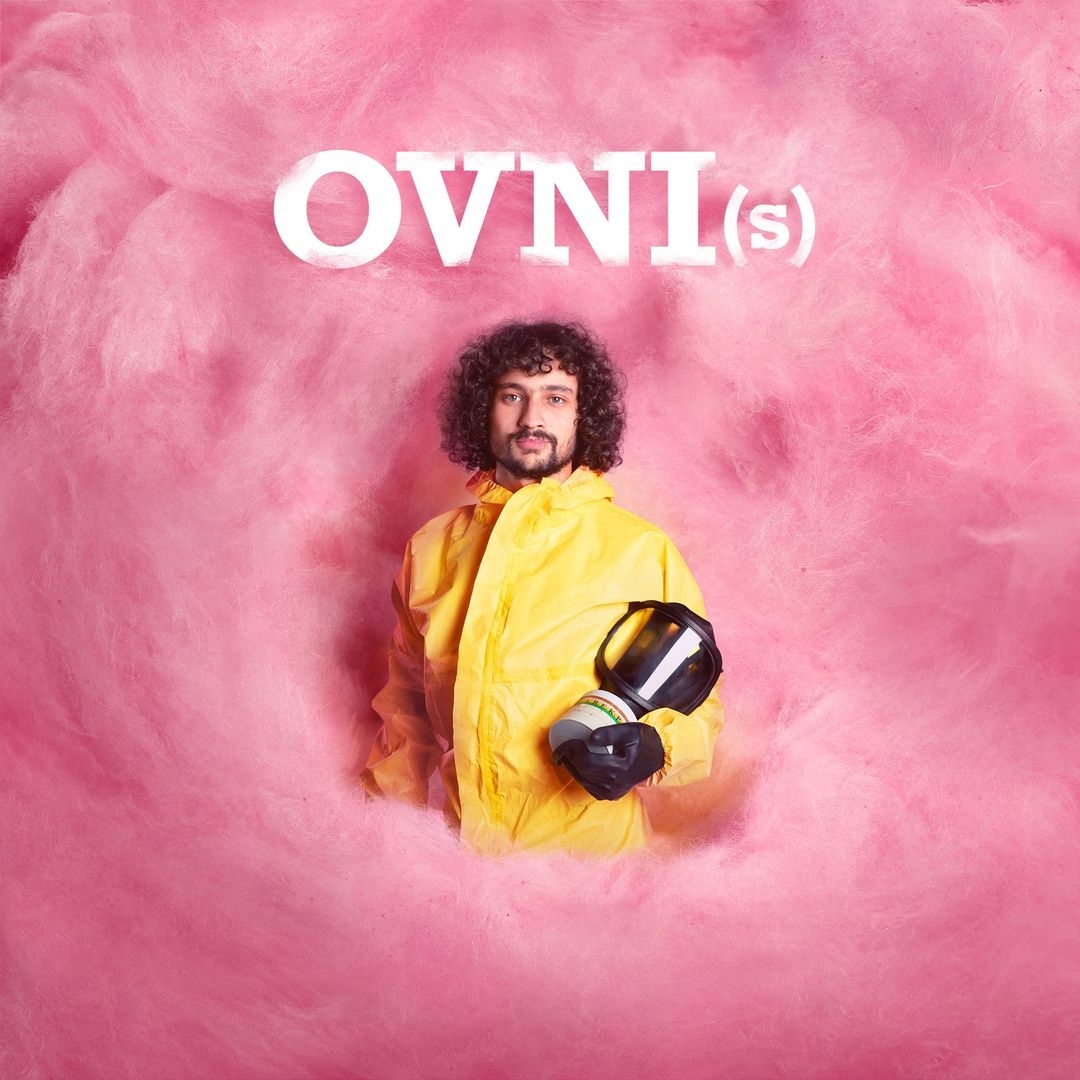 Man in yellow hazmat suit holds gas mask against pink cotton candy background. Studio shot, eye-level angle, surreal. OVNI(s) text visible.