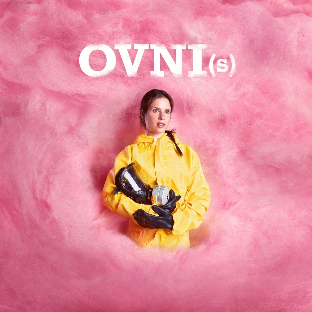 Woman in yellow hazmat suit holds gas mask against a pink cotton candy background with the text “OVNI(s)” above her head.