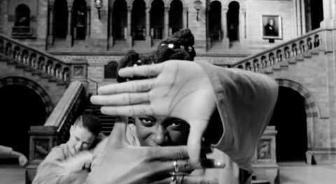 Monochrome shot of a person framing with hands in an ornate interior, another person blurred in the background, enhancing depth.