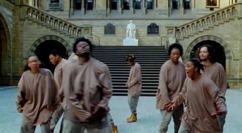 Group of people in beige outfits perform dance in grand hall with arched entryways, staircase, and statue; bright, symmetrical.