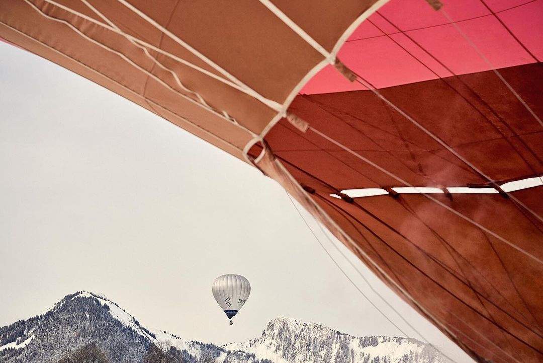 Low-angle view of a multicolored hot air balloon canopy and a white hot air balloon in flight over snow-capped mountains against a pale sky.