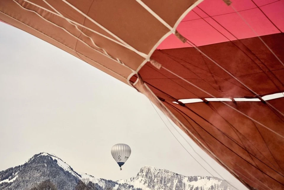 Low-angle view of a multicolored hot air balloon canopy and a white hot air balloon in flight over snow-capped mountains against a pale sky.