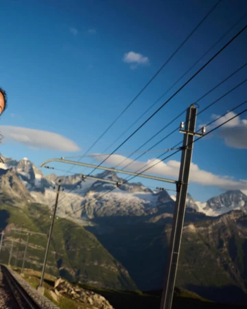 Scenic mountain landscape featuring a railway track and power lines under a blue sky, with visible mountain peaks.