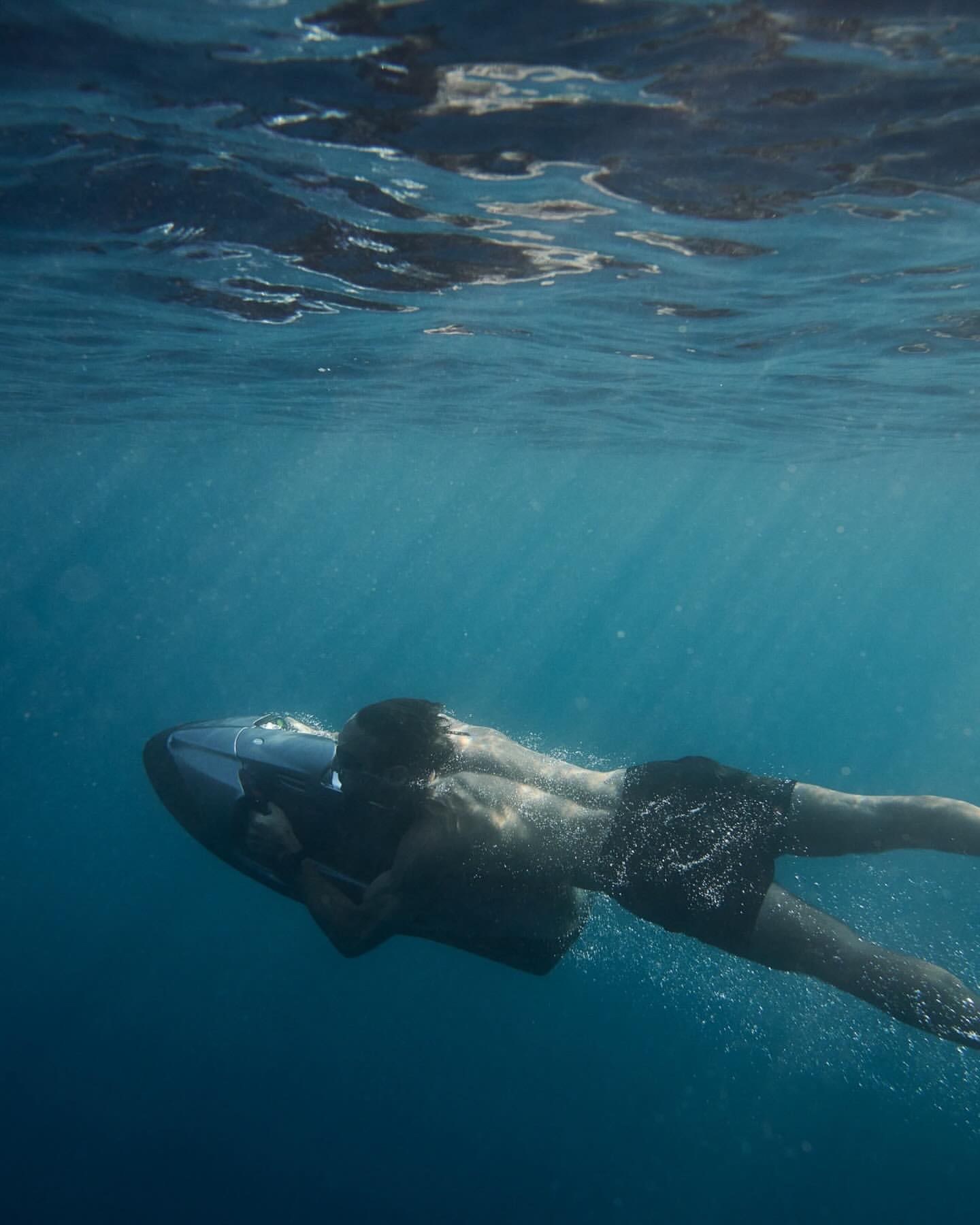 Underwater view of a man swimming with a personal watercraft in the ocean, wearing black shorts, showcasing the ocean environment.
