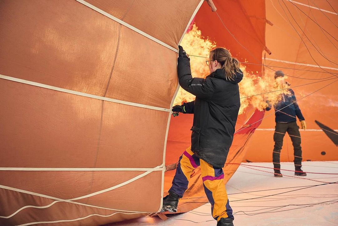 Two people prepare a hot air balloon for flight, one holding open the fabric as flames fill the balloon on a snowy outdoor field.