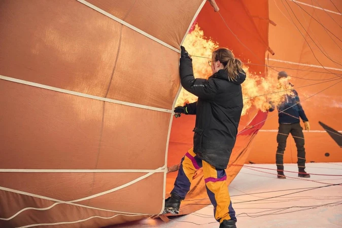 Two people prepare a hot air balloon for flight, one holding open the fabric as flames fill the balloon on a snowy outdoor field.