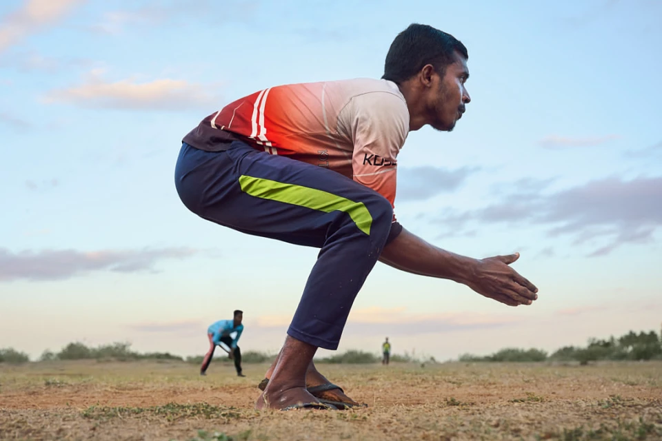 A cricket bowler in action, preparing to deliver the ball on a dirt field under a cloudy sky, with other players visible in the background.