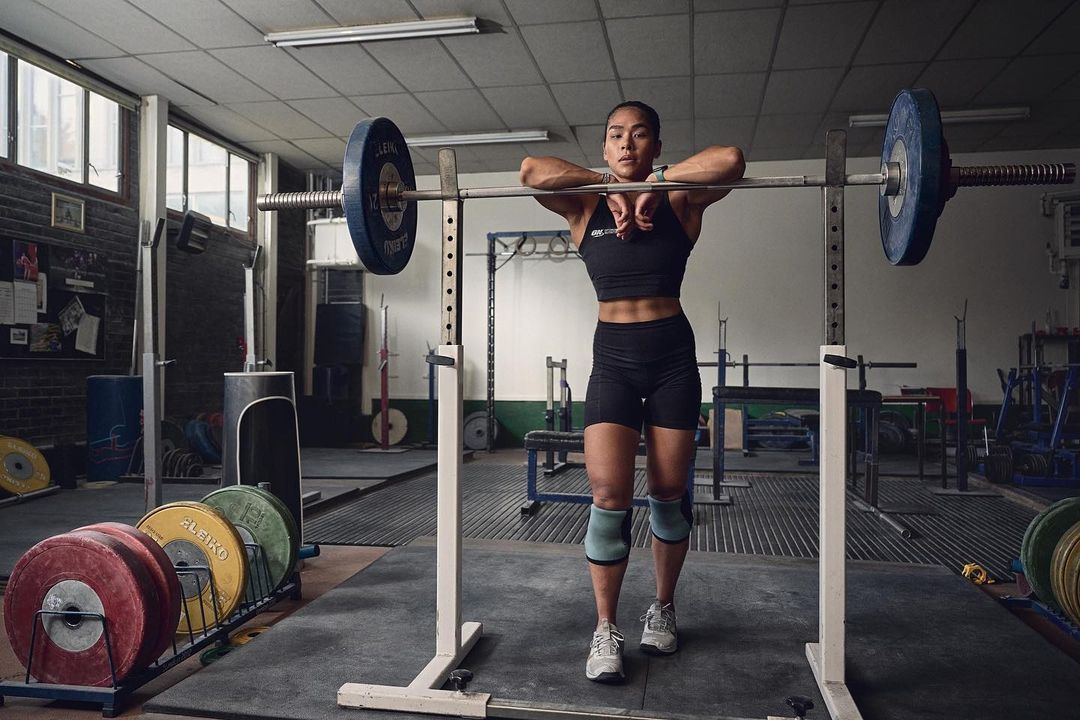 Female weightlifter with barbell in gym. Adult woman resting on barbell. Weight plates, weights, gym equipment in the background.