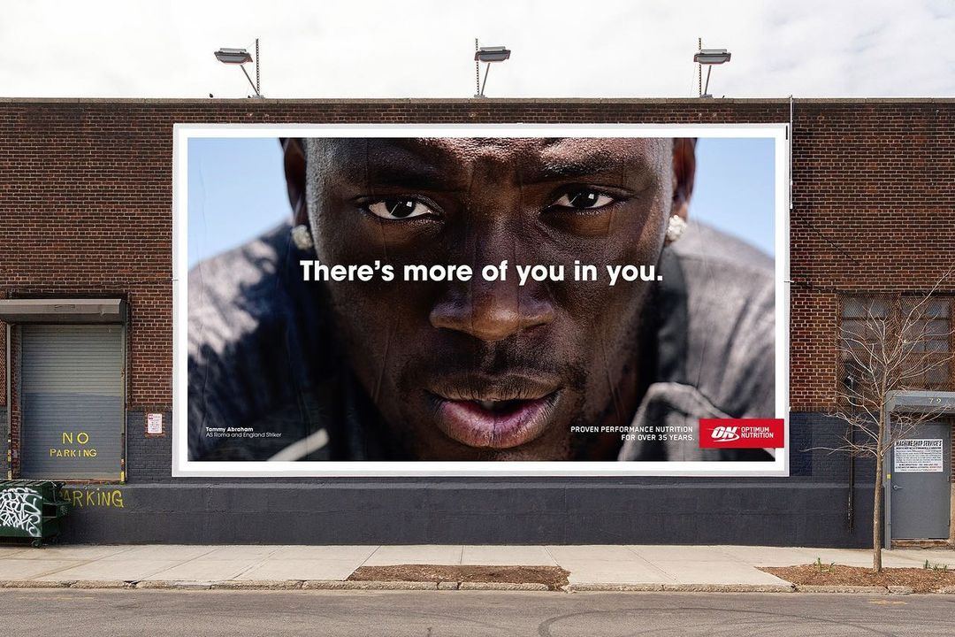 Close-up billboard of Tommy Abraham, England Striker, with tagline "There's more of you in you," against a brick wall in urban setting.