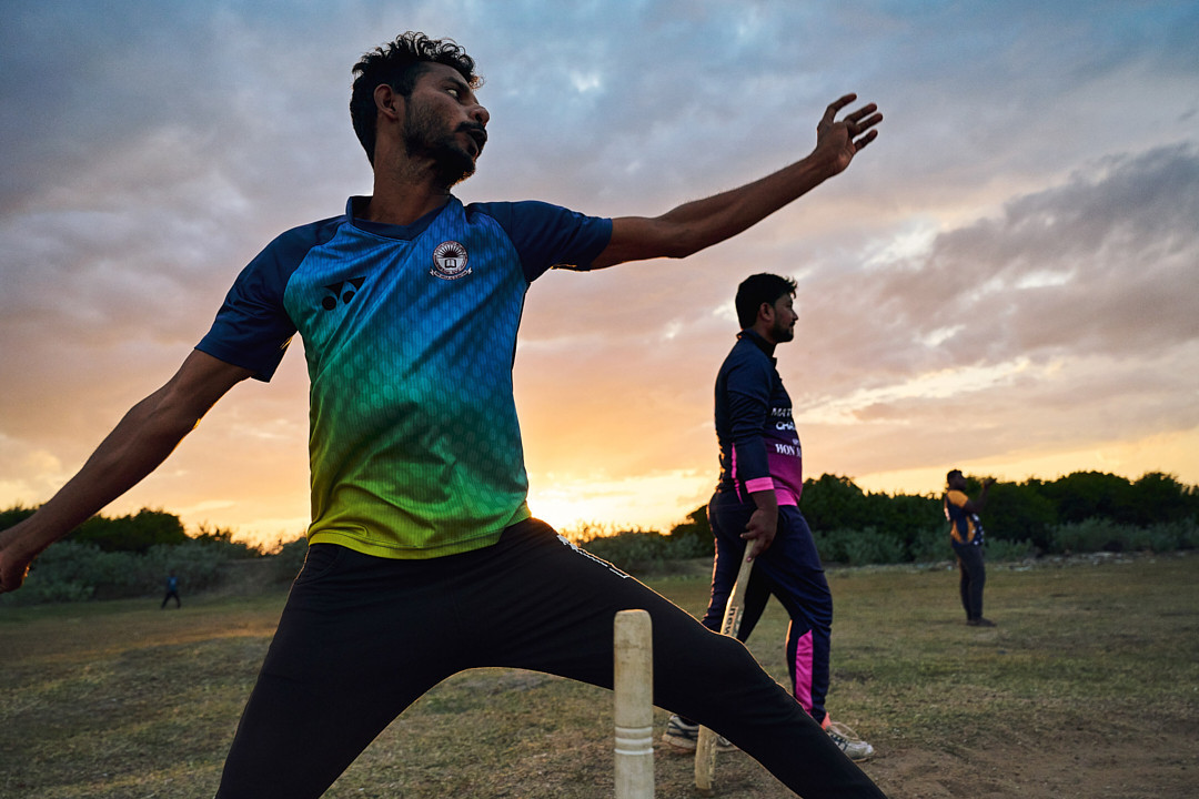 A cricket bowler in action on a field at dusk, with teammates in the background, captured in a wide shot with a cloudy sky.