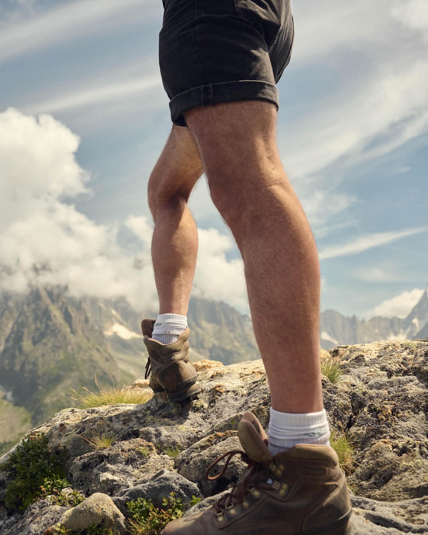 Hiker's legs and hiking boots ascend a rocky mountain trail, set against a backdrop of sky and distant mountain peaks.
