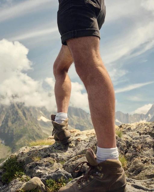 Hiker's legs and hiking boots ascend a rocky mountain trail, set against a backdrop of sky and distant mountain peaks.