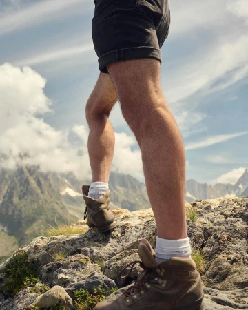 Hiker's legs and hiking boots ascend a rocky mountain trail, set against a backdrop of sky and distant mountain peaks.