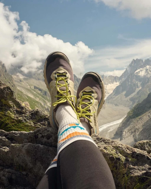 Low angle shot of hiking shoes and striped socks with scenic view of rocky mountain peaks and cloudy blue sky in the background.