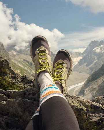 Low angle shot of hiking shoes and striped socks with scenic view of rocky mountain peaks and cloudy blue sky in the background.