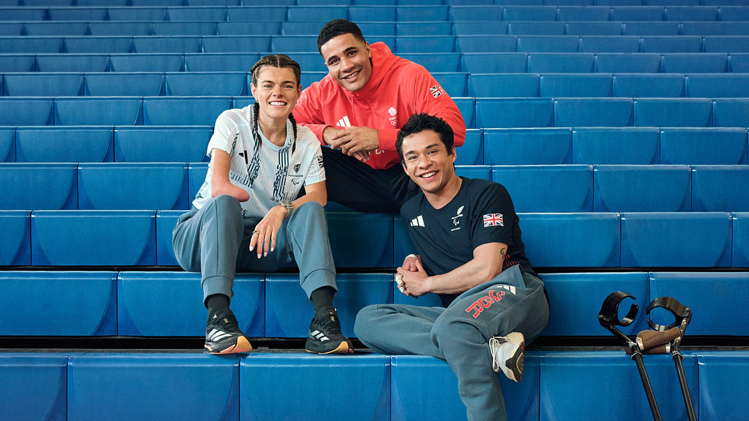 Group photo of three smiling athletes with disabilities seated on blue stadium steps, wearing Adidas apparel, showcasing sports inclusivity.