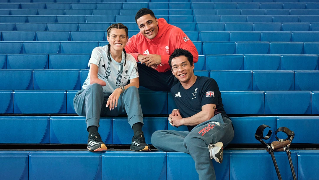Group photo of three smiling athletes with disabilities seated on blue stadium steps, wearing Adidas apparel, showcasing sports inclusivity.