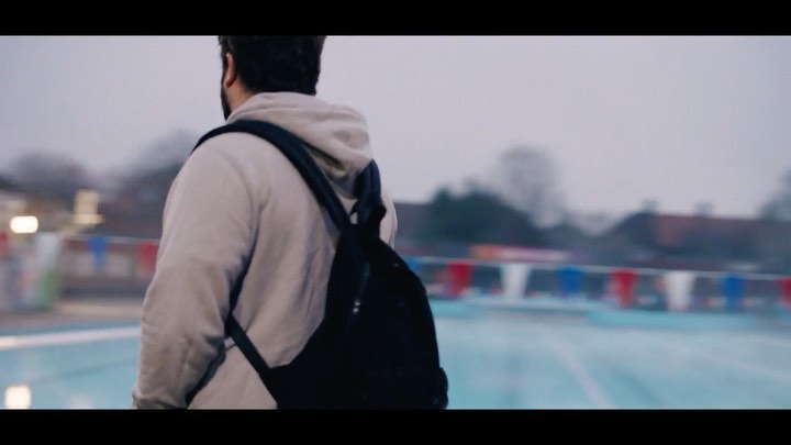 Adult male with black backpack walks by an outdoor swimming pool on a cloudy day in medium shot, back view, muted colors.
