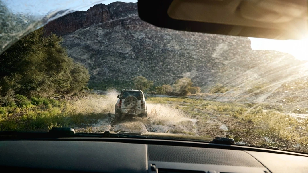 Off-road vehicle driving through a shallow river with a mountain backdrop, captured from the interior of another car on a sunny day.