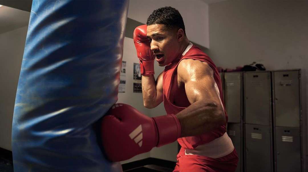Sweaty boxer in red tank top and gloves throwing a punch at a blue heavy bag in a gym with lockers visible in the background.