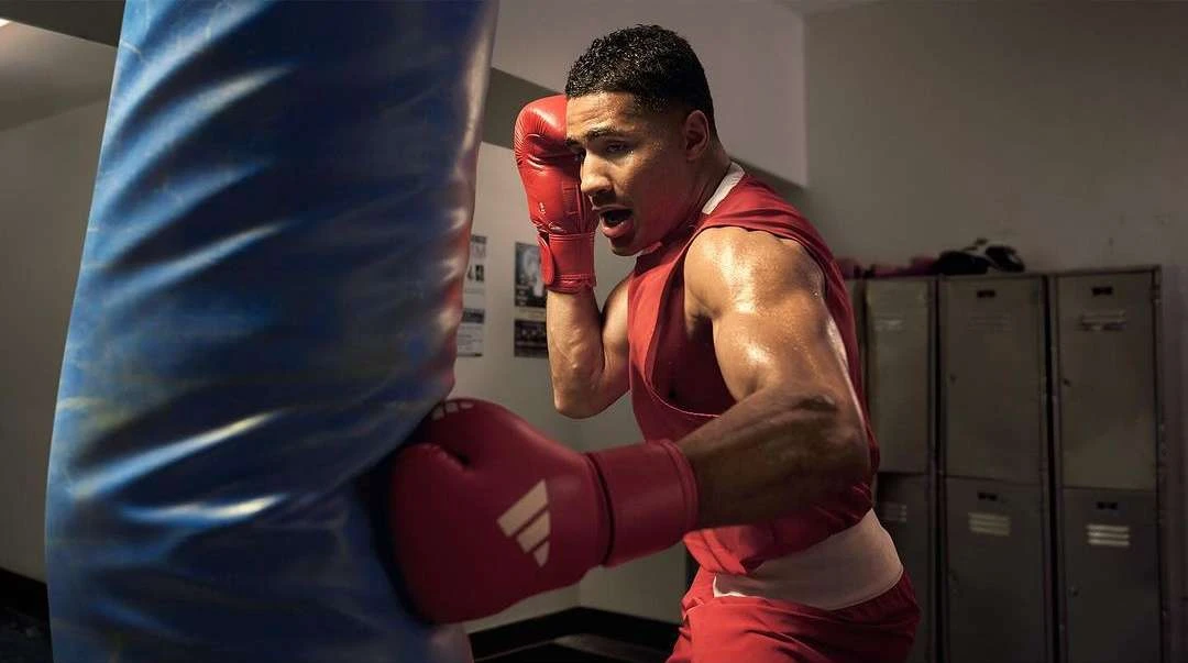 Sweaty boxer in red tank top and gloves throwing a punch at a blue heavy bag in a gym with lockers visible in the background.