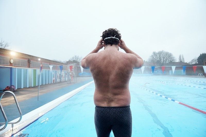 Athletic man wearing swim goggles standing by an outdoor swimming pool with blue water on an overcast day. Fitness and outdoor sports.