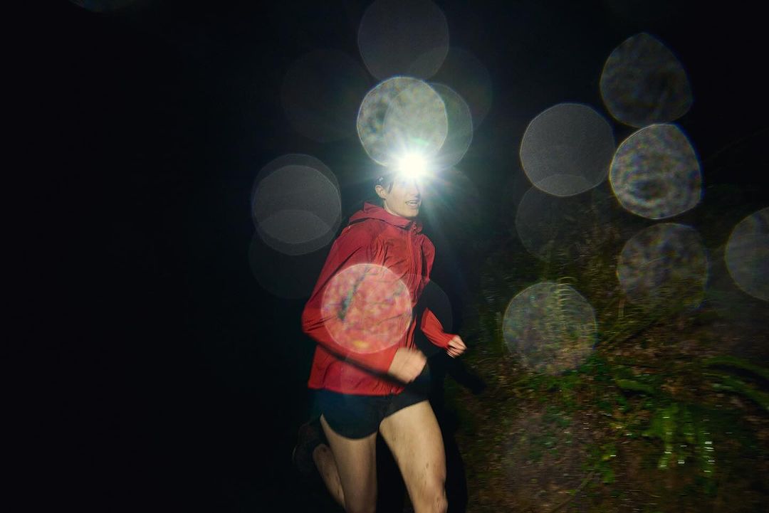 Woman runs on a dark trail at night with a headlamp. She wears a red jacket, athletic shorts, and running shoes in an outdoor setting.