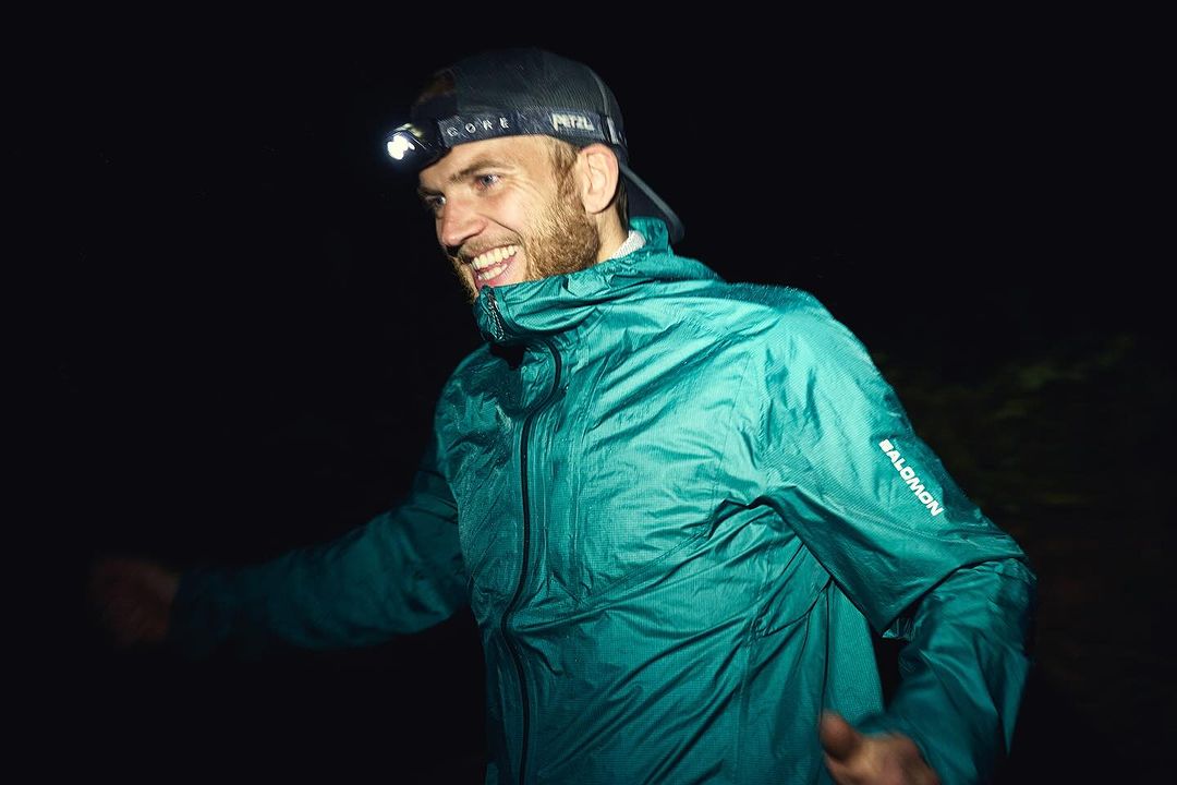 Smiling adult man in running gear and headlamp outdoors at night. He is wearing a teal hooded jacket with a Salomon logo on the sleeve.