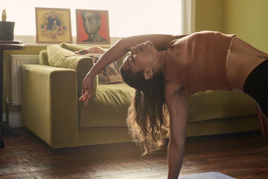 A woman in a brown shirt and black leggings does a backbend in a living room with a sofa, art prints, and natural light.