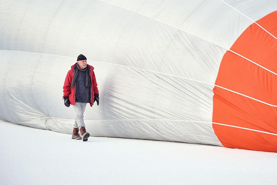 Man in red jacket walks beside a large partially inflated hot air balloon in a snowy field, creating a winter scene.