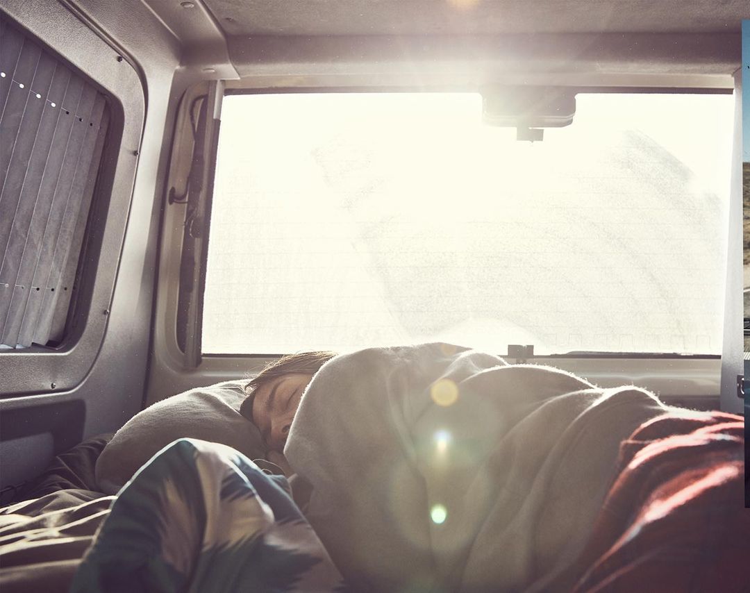 Person sleeping in the back of a vehicle with window covers. Interior view shows bedding and head resting on a pillow. Travel lifestyle.