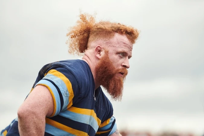 Profile portrait of a white man with red hair in a ponytail and a thick red beard wearing a striped rugby jersey.