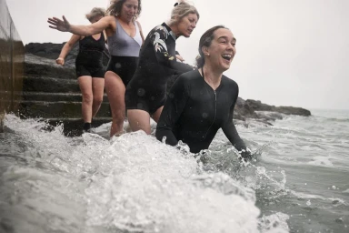 Four women in swimwear emerging from ocean water onto rocky shore, photographed by Sam Bénard in London.